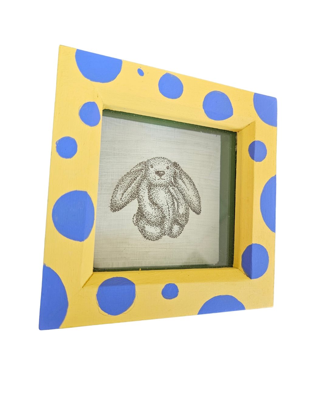 A square decorative picture frame with a yellow border and blue polka dots, holding a sketch of a rabbit on textured paper, shown against a plain white background.