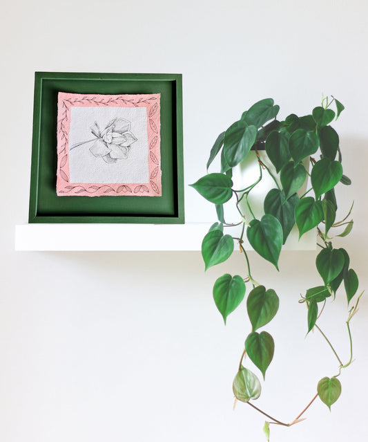 Framed artwork of a flower on a shelf with a green plant.