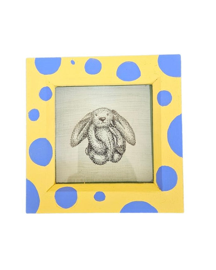 A square decorative picture frame with a yellow border patterned with blue polka dots, holding a small sketch of a bunny on a light background.