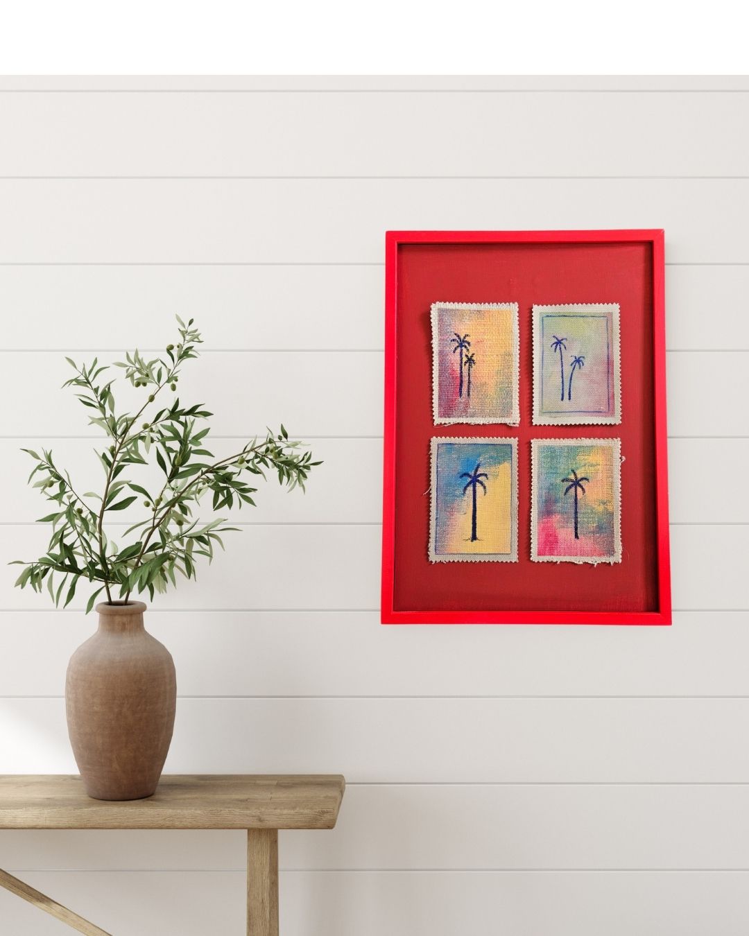 A red shadowbox frame displaying four colorful palm tree paintings, mounted on a white shiplap wall above a wooden table with a brown vase of green leaves.