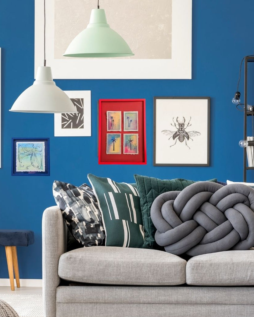 A styled living room scene featuring a light gray fabric sofa with decorative throw pillows, including a large gray knotted pillow. The wall behind the sofa is a vibrant blue, decorated with framed art, two hanging pendant lights, and a black shelving unit.