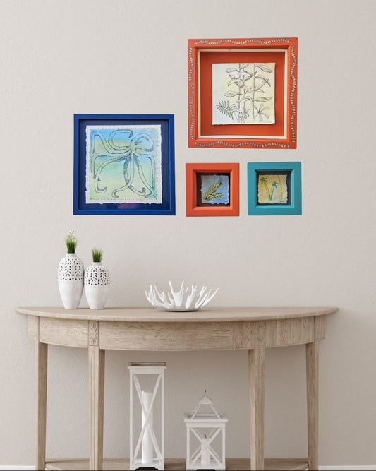 A styled interior scene showing four framed wall art pieces in blue, orange, and teal frames hanging above a light wood console table with decorative vases and lanterns.