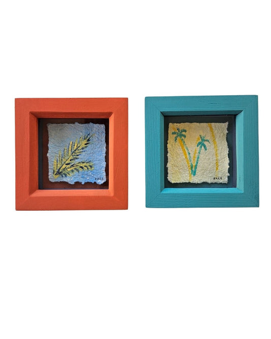Two square framed art pieces on a white background. The left frame is coral orange and contains a textured blue square with a yellow palm leaf.